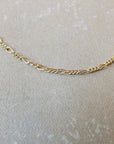 A close-up view of the Figaro Chain Necklace by Becoming Jewelry, featuring a delicate and intricate design, laid on a textured beige surface, showcasing its refined craftsmanship.