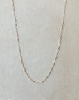 A delicate, thin Figaro Chain Necklace from Becoming Jewelry is displayed against a plain beige background, enhancing its intricate texture with a layered look.