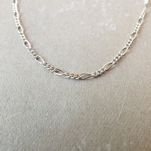 A close-up of a sterling silver Becoming Jewelry Figaro chain necklace on a textured surface.