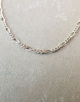 The Figaro Chain Necklace by Becoming Jewelry, featuring a delicate gold chain with a unique Figaro link pattern and layered look, is laid out on a light gray surface.