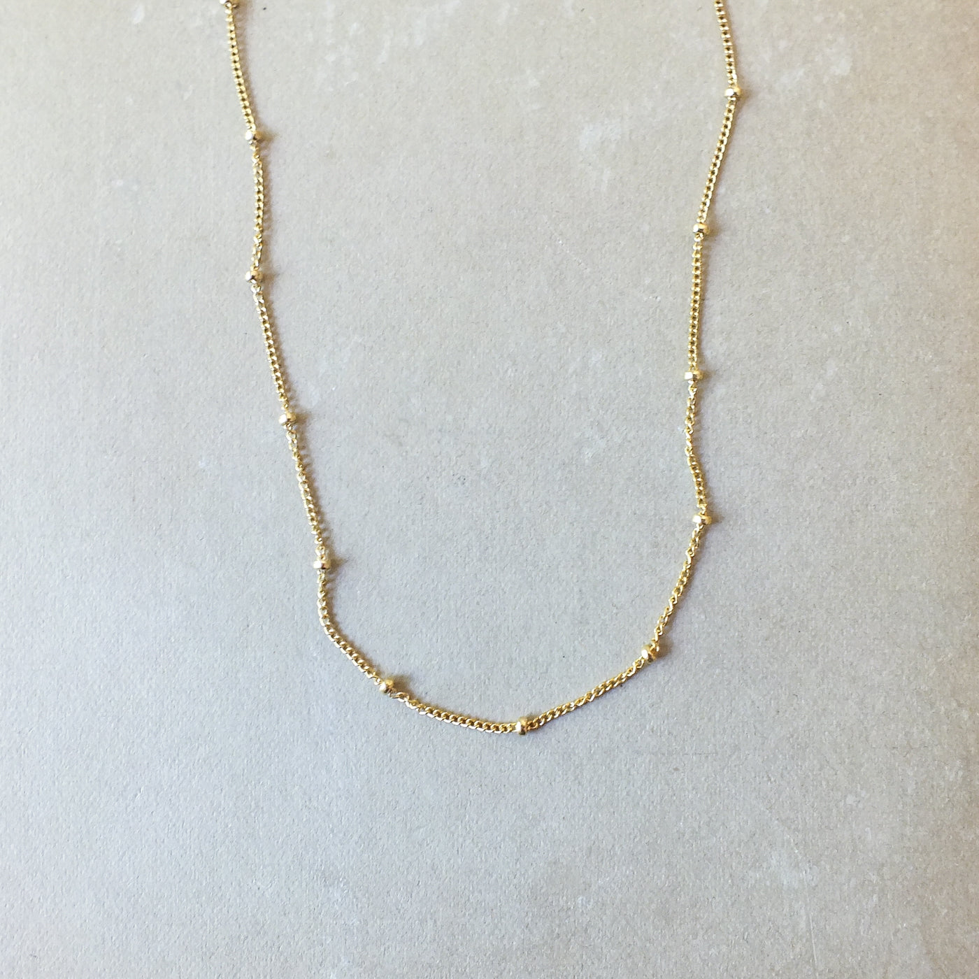 The Satellite Chain Necklace by Becoming Jewelry is a delicate gold satellite chain adorned with small beads evenly spaced along its length, elegantly displayed on a light gray surface. Perfect for layering with other necklaces to achieve a stylish, everyday look.