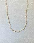 The Satellite Chain Necklace by Becoming Jewelry is a delicate gold satellite chain adorned with small beads evenly spaced along its length, elegantly displayed on a light gray surface. Perfect for layering with other necklaces to achieve a stylish, everyday look.