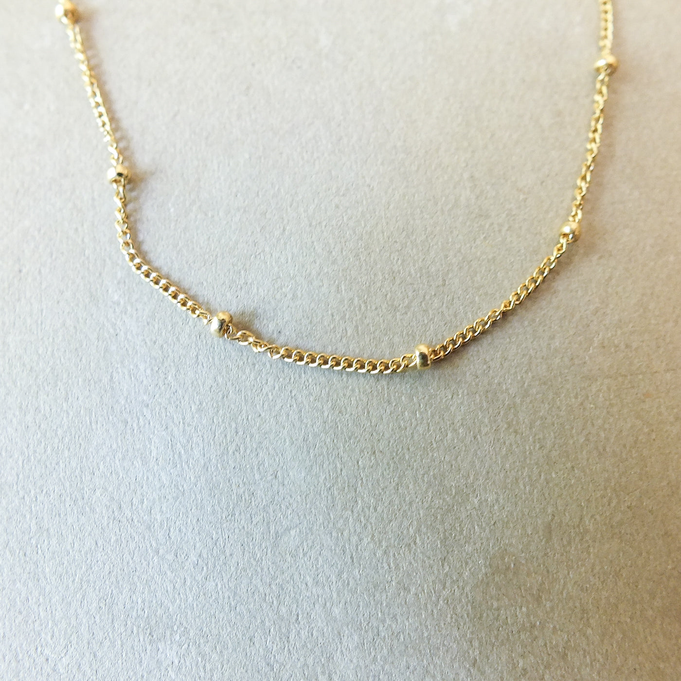 Presenting the Becoming Jewelry Satellite Chain Necklace, adorned with small, evenly spaced beads, elegantly presented on a beige surface. It's an ideal piece for layering to achieve a graceful style or wearing solo as your everyday go-to necklace.