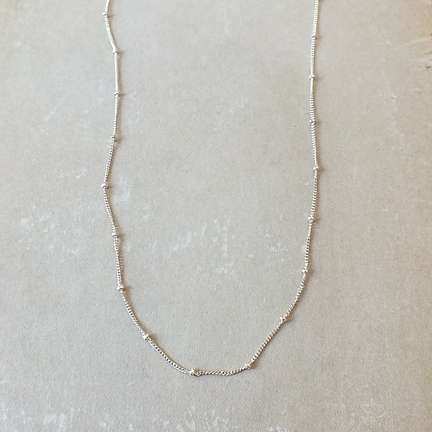 A delicate, thin Satellite Chain Necklace from Becoming Jewelry rests on a light-colored surface, ideal for layering with other necklaces to achieve an everyday look.
