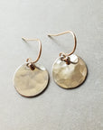 The Hammered Disc Drop Earrings, small by Becoming Jewelry feature gold, hand-hammered round discs on a light gray background with French hook ear wires.