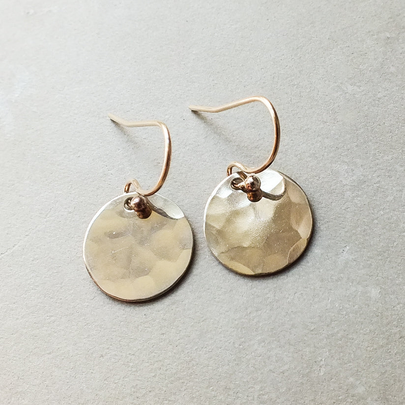 A pair of small, Becoming Jewelry Hammered Disc Drop Earrings on a grey surface.