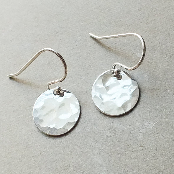 A pair of small Hammered Disc Drop Earrings by Becoming Jewelry, featuring round, hand-hammered silver discs attached to simple french hook ear wires, is set against a neutral background.
