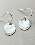 A pair of small Hammered Disc Drop Earrings by Becoming Jewelry, featuring round, hand-hammered silver discs attached to simple french hook ear wires, is set against a neutral background.