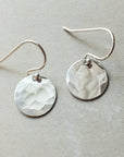 The Hammered Disc Drop Earrings, small by Becoming Jewelry are a pair of silver-toned, lightweight earrings featuring hand-hammered discs and French hook ear wires, displayed on a light gray background.