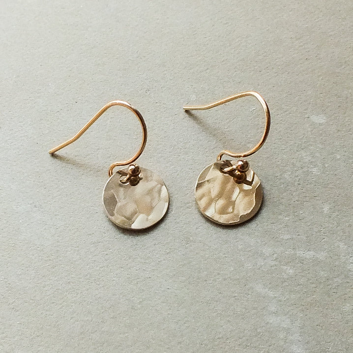 A pair of tiny Hammered Disc Drop Earrings by Becoming Jewelry, featuring lightweight, hand-hammered gold discs with hook fastenings, displayed on a gray surface—perfect for everyday wear.