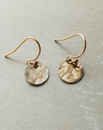 A pair of tiny Hammered Disc Drop Earrings by Becoming Jewelry, featuring lightweight, hand-hammered gold discs with hook fastenings, displayed on a gray surface—perfect for everyday wear.