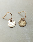 A pair of Becoming Jewelry hammered gold filled tiny disc drop earrings on a neutral surface.