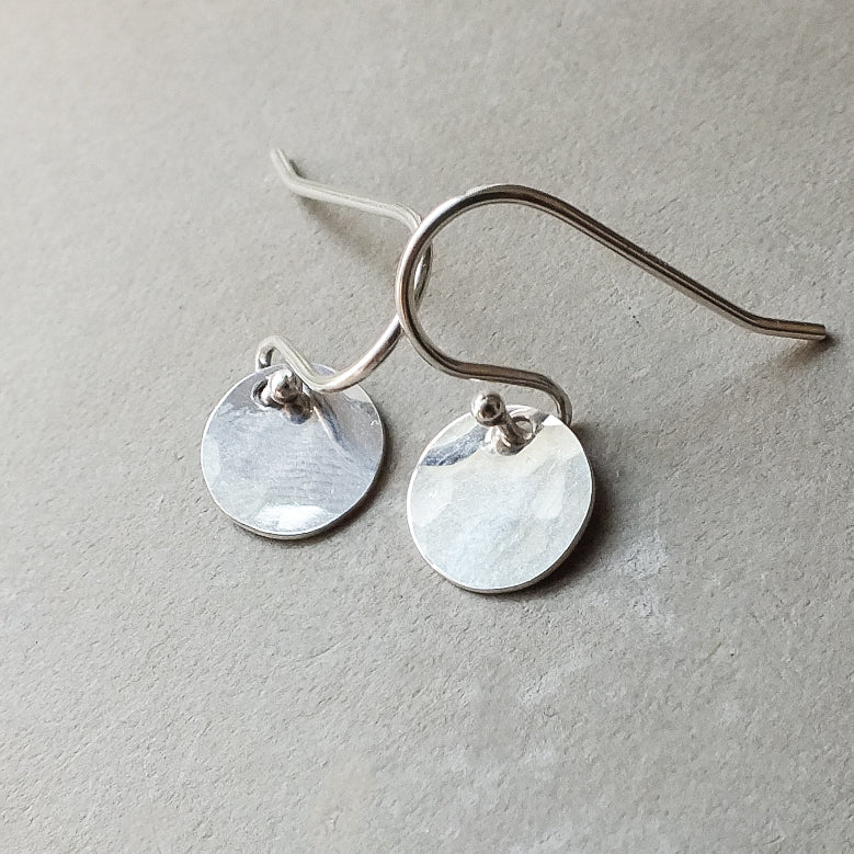 Introducing the Hammered Disc Drop Earrings, tiny by Becoming Jewelry—exquisite silver hook earrings with lightweight, hand-hammered discs, elegantly displayed on a gray surface and perfect for everyday wear.