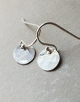 Introducing the Hammered Disc Drop Earrings, tiny by Becoming Jewelry—exquisite silver hook earrings with lightweight, hand-hammered discs, elegantly displayed on a gray surface and perfect for everyday wear.