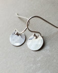 A pair of tiny Becoming Jewelry Hammered Disc Drop Earrings on a gray surface.