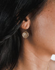 Close-up of a person wearing Becoming Jewelry's Hammered Disc Drop Earrings, small, featuring a round, hand-hammered pendant. The earrings dangle elegantly from the earlobe, secured with delicate French hook ear wires. The person's dark hair is partially visible in the shot.
