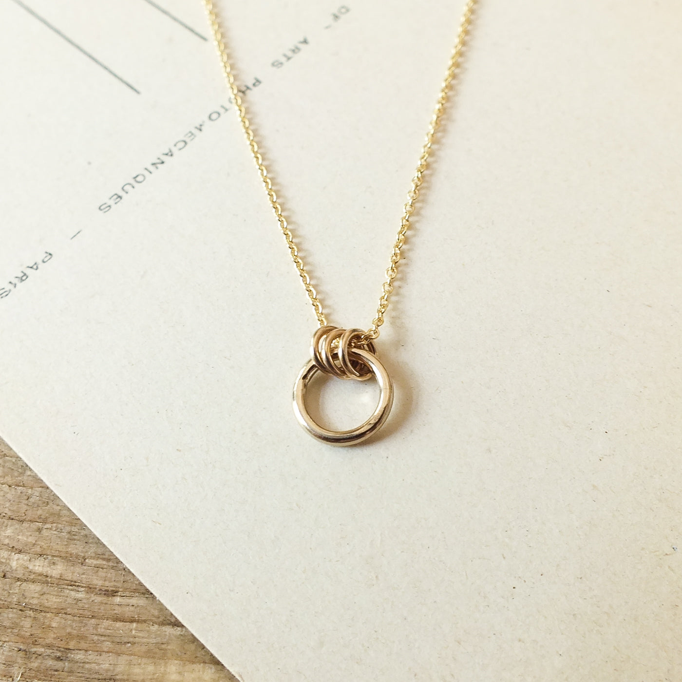 Close-up of the exquisite Family Circle Necklace by Becoming Jewelry, showcasing a pendant with three interlocking rings symbolizing strength and love, displayed on a textured white surface. This beautiful piece makes the perfect go-to gift.