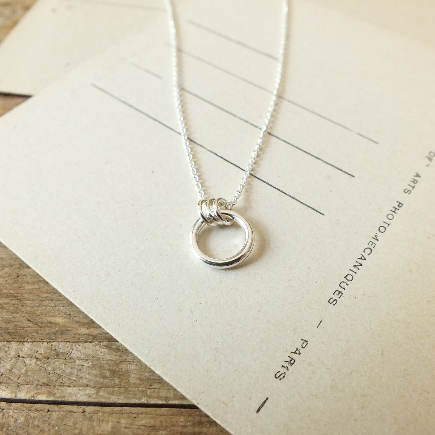A silver Family Circle Necklace by Becoming Jewelry, featuring a circular pendant, lies on a beige piece of paper on a wooden surface, symbolizing strength and love.
