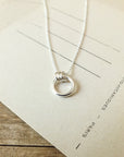 A silver Family Circle Necklace by Becoming Jewelry, featuring a circular pendant, lies on a beige piece of paper on a wooden surface, symbolizing strength and love.