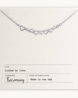 The Heart Links Necklace from Becoming Jewelry features silver heart links on a "Linked by Love" white card, marked "Made in the USA." This elegant chain embodies love's essence through its graceful design.