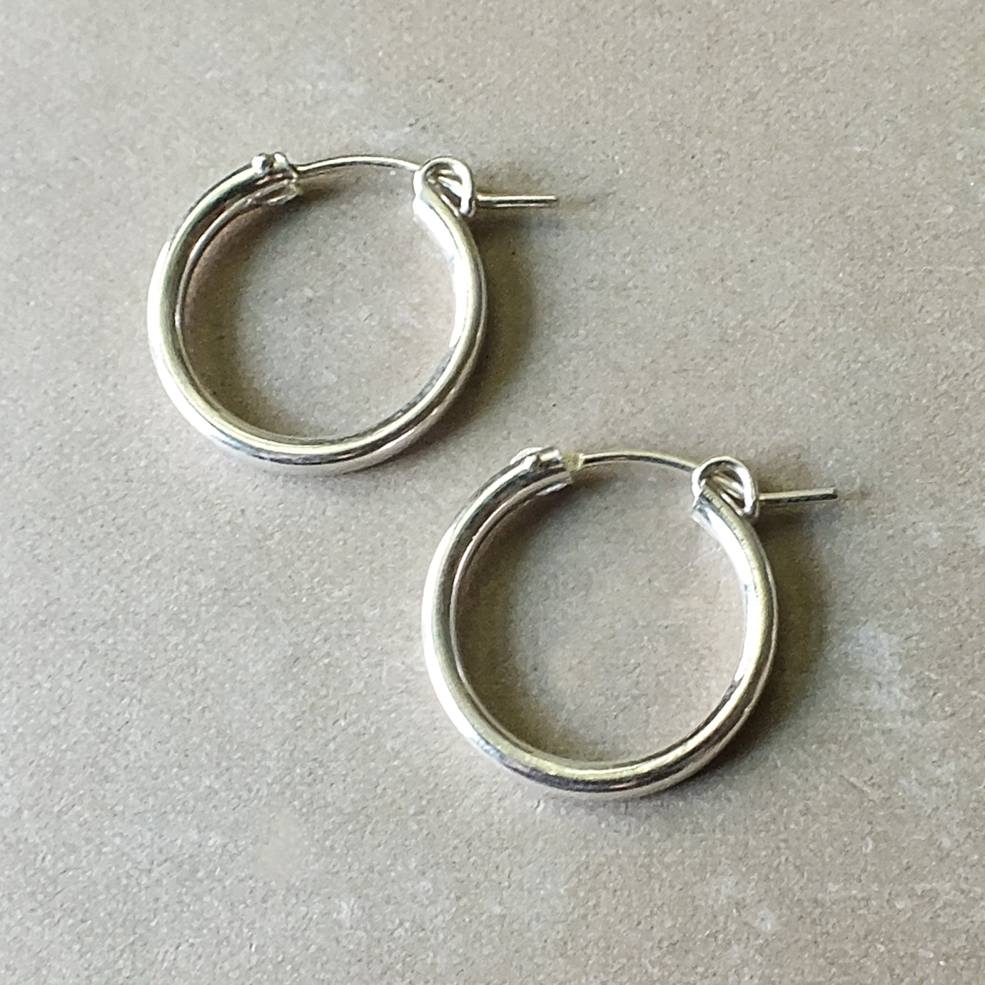 Two large silver hoop earrings from Becoming Jewelry, perfect for everyday wear, are elegantly placed on a beige surface.