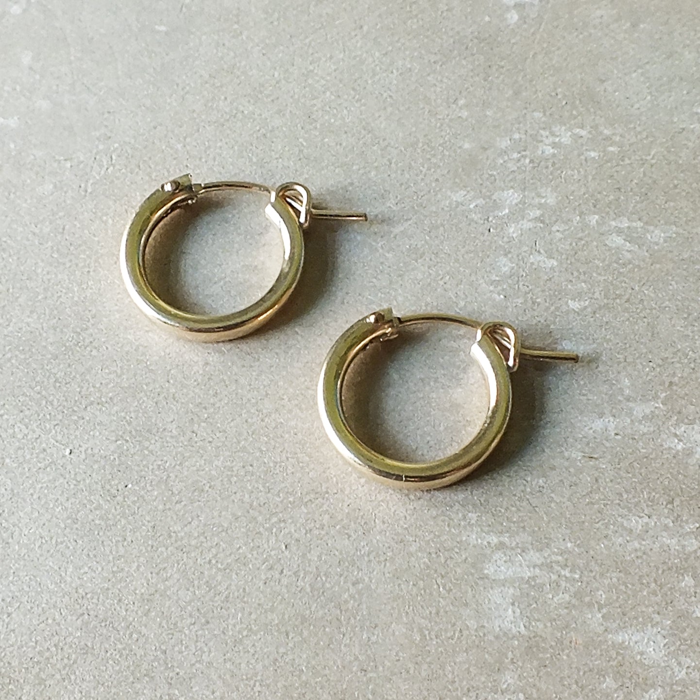 A pair of medium-sized Everyday Hoop Earrings in gold, classic and comfortable for any occasion, from Becoming Jewelry, placed on a light gray, textured surface.
