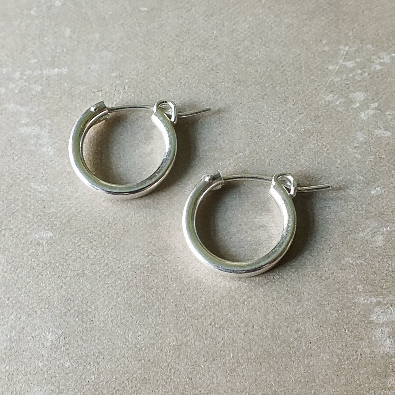A pair of medium, silver hoop earrings with hinge clasps placed on a light beige surface. These classic Everyday Hoop Earrings from Becoming Jewelry offer a comfortable fit perfect for any occasion.