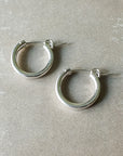 A pair of medium, silver hoop earrings with hinge clasps placed on a light beige surface. These classic Everyday Hoop Earrings from Becoming Jewelry offer a comfortable fit perfect for any occasion.