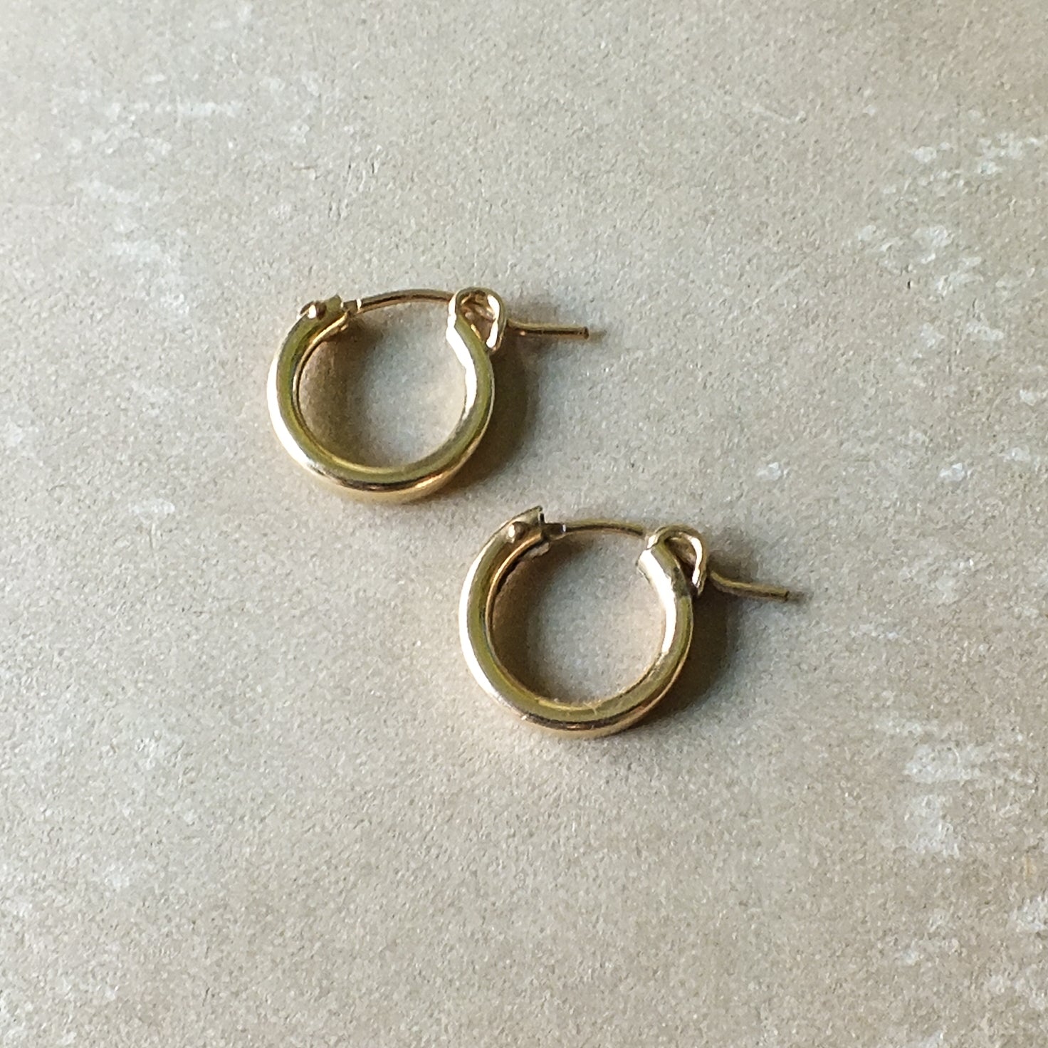 A pair of Becoming Jewelry's Everyday Hoop Earrings, in a small size and classic lightweight gold design, lying on a beige surface—perfect for everyday wear.
