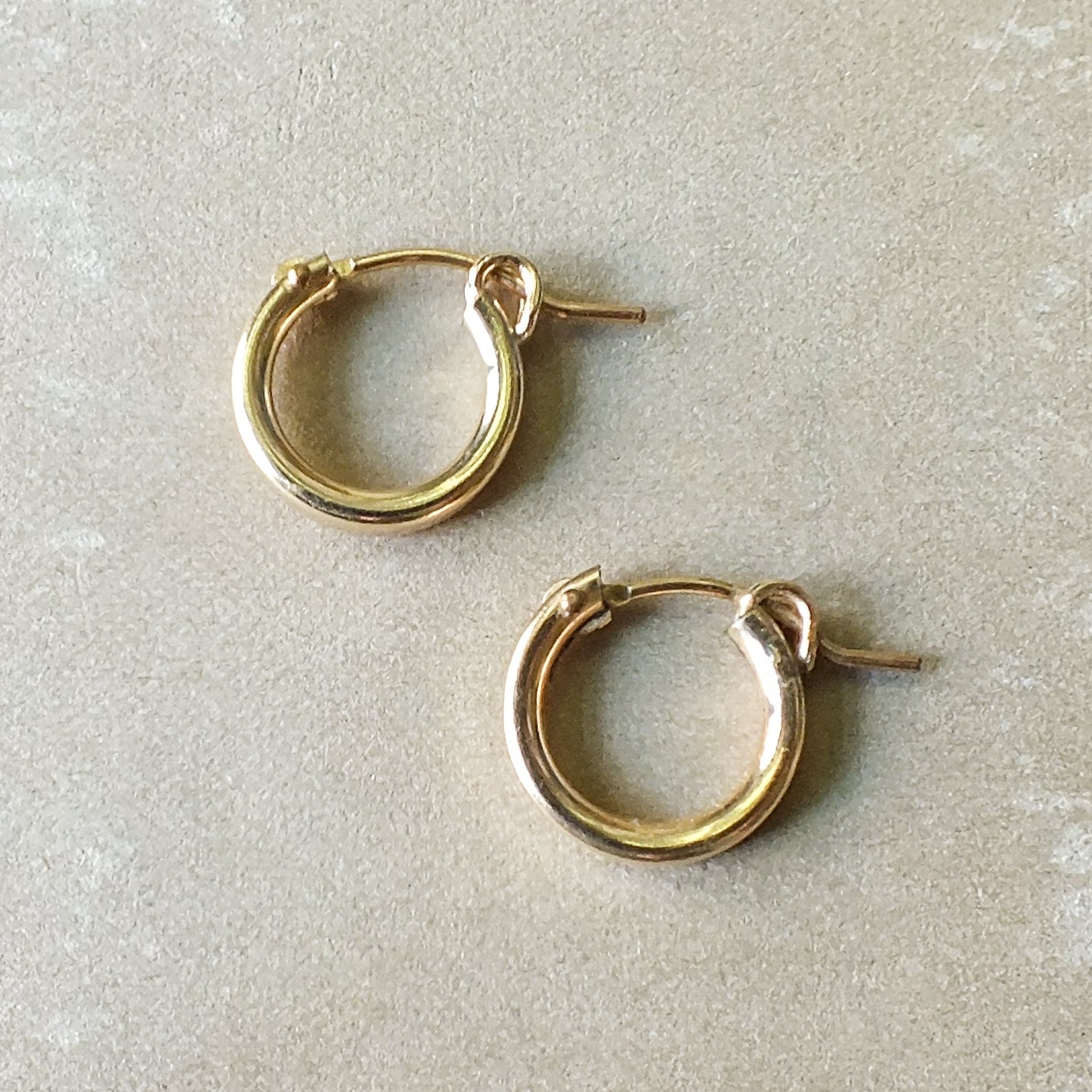 A pair of Becoming Jewelry's Everyday Hoop Earrings, small, lying on a light grey surface perfect for everyday wear.