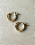 A pair of Becoming Jewelry's Everyday Hoop Earrings, in a small size and classic lightweight gold design, lying on a beige surface—perfect for everyday wear.
