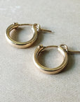 A pair of small Everyday Hoop Earrings by Becoming Jewelry, made of classic gold and featuring latch-back closures, lies on a light beige surface, offering a lightweight touch.