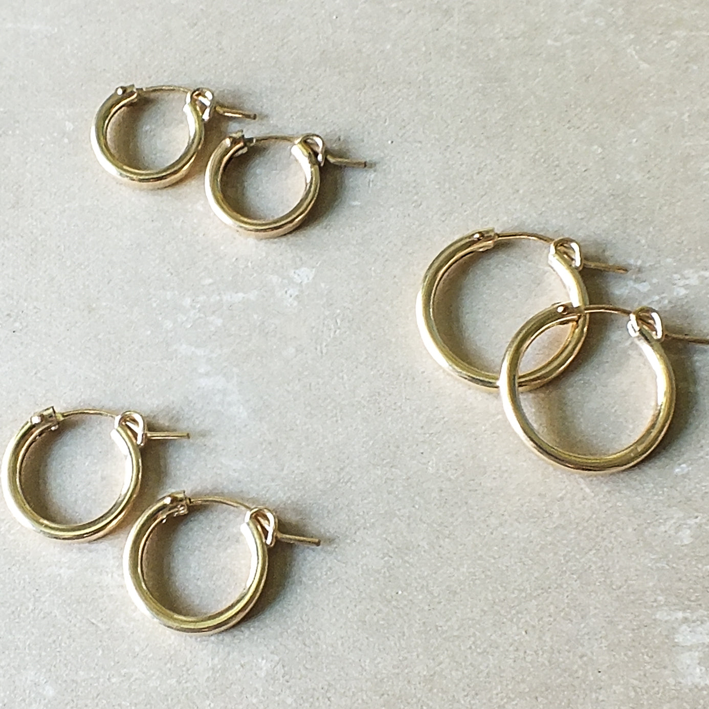 A collection of Becoming Jewelry&#39;s medium Everyday Hoop Earrings in gold filled on a gray surface.