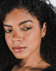 A close-up of a person with curly dark hair, wearing Becoming Jewelry's Everyday Hoop Earrings (small) and a necklace, looking slightly to the side with a neutral expression.