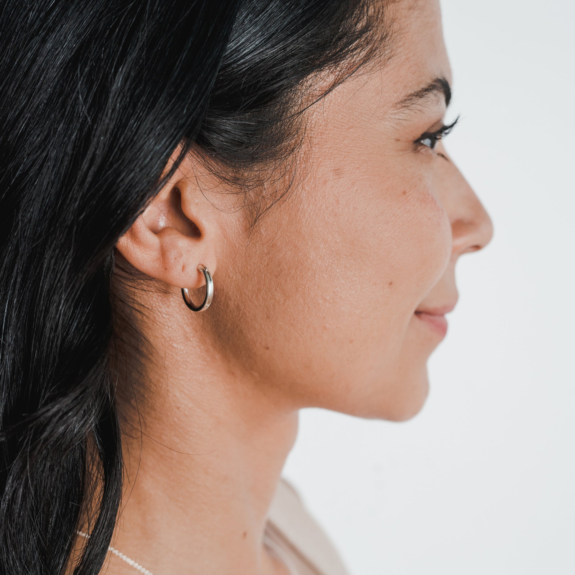 Profile view of a person with long dark hair, wearing a medium-sized, classic Everyday Hoop Earring by Becoming Jewelry in their left ear. The background is plain white.
