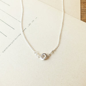 Becoming Jewelry's Love Knot Necklace with a swirl pendant on a wooden surface.