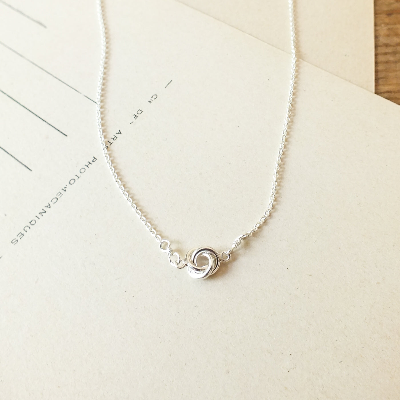 Becoming Jewelry&#39;s Love Knot Necklace with a swirl pendant on a wooden surface.