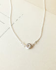 A delicate Love Knot Necklace from Becoming Jewelry, featuring a small intertwined knot pendant on a silver chain, is elegantly placed on a beige paper background.