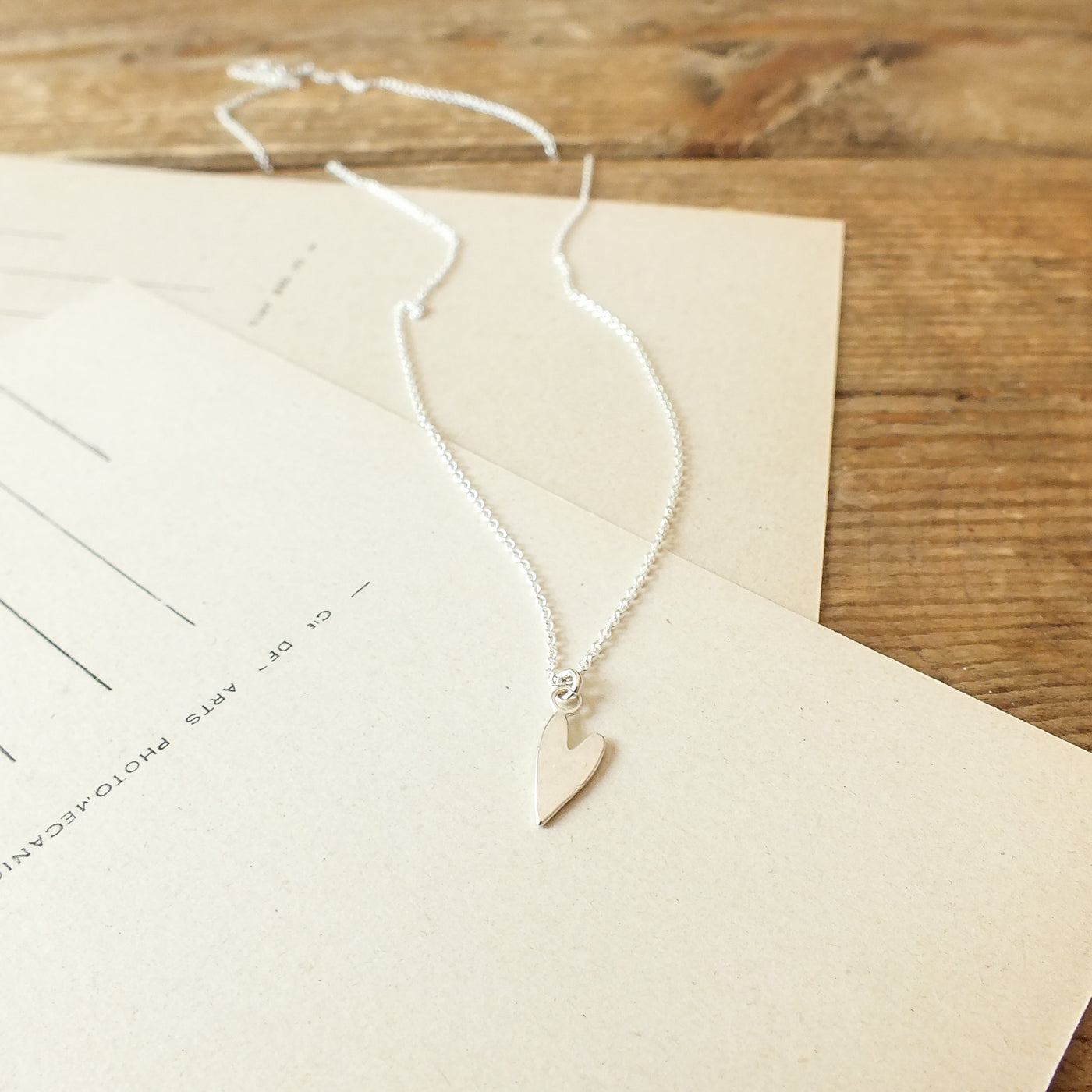 A delicate Love Deeply necklace from Becoming Jewelry, featuring a silver heart-shaped pendant, rests on a beige envelope atop a wooden surface.