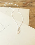 A delicate Love Deeply necklace from Becoming Jewelry, featuring a silver heart-shaped pendant, rests on a beige envelope atop a wooden surface.