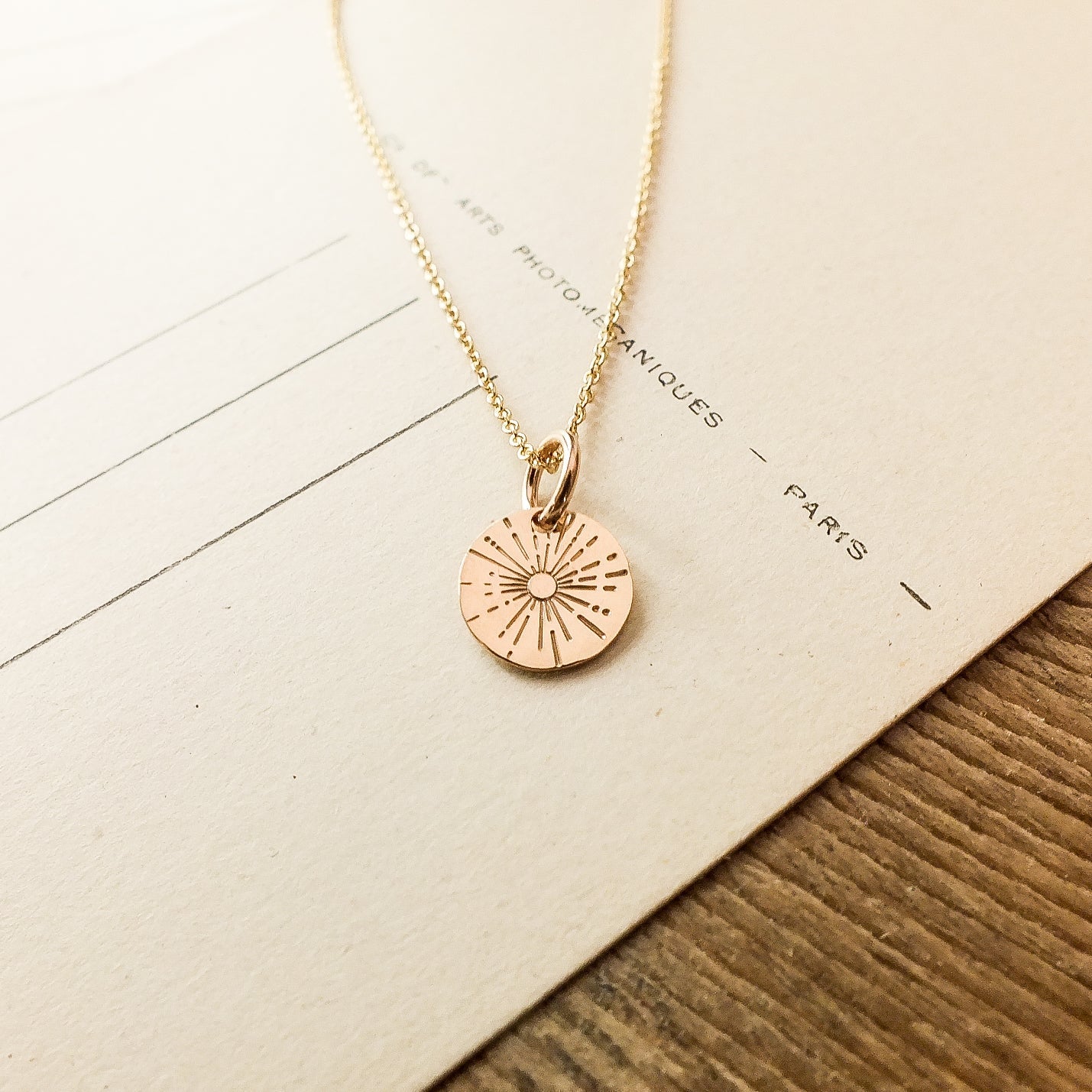 A gold "Be The Light Necklace" by Becoming Jewelry, featuring a circular pendant with radiant sunburst beams, elegantly placed on beige paper with faint writing.