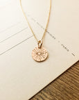A gold "Be The Light Necklace" by Becoming Jewelry, featuring a circular pendant with radiant sunburst beams, elegantly placed on beige paper with faint writing.