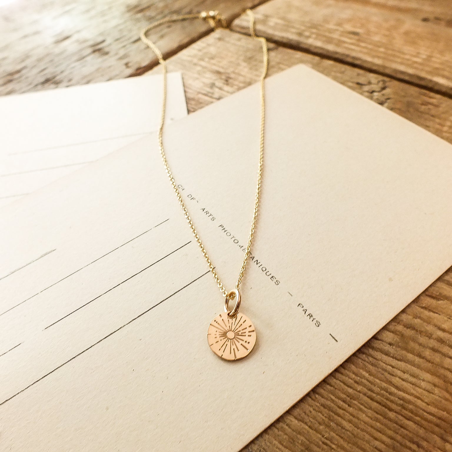 The Be The Light Necklace from Becoming Jewelry, featuring a circular sunburst pendant with radiant beams, lies on a wooden surface atop postcards or lined paper.