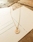 The Be The Light Necklace from Becoming Jewelry, featuring a circular sunburst pendant with radiant beams, lies on a wooden surface atop postcards or lined paper.