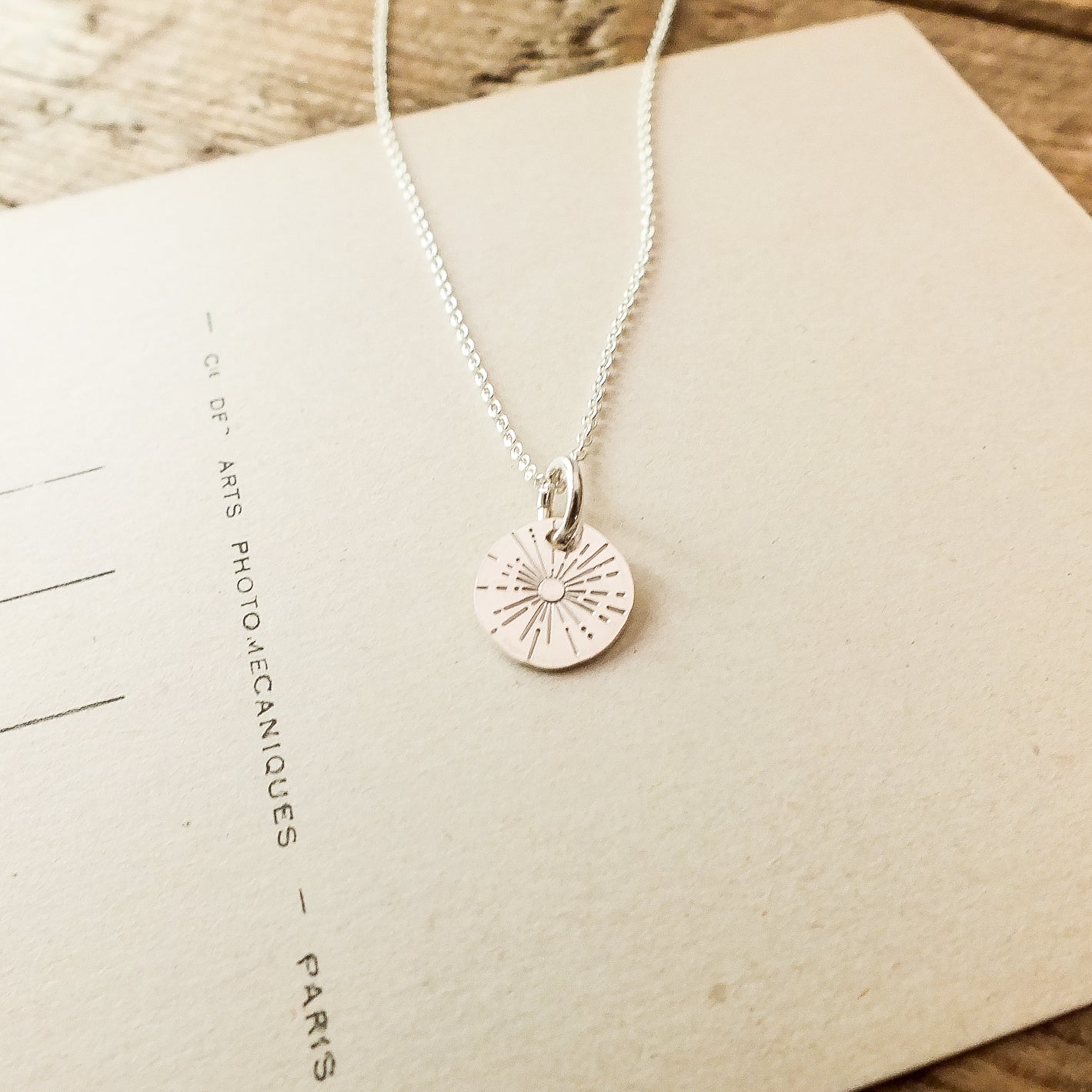 The Be The Light Necklace by Becoming Jewelry, featuring a silver charm engraved with a sunburst design, is placed on a piece of paper with text.