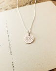 The Be The Light Necklace by Becoming Jewelry, featuring a silver charm engraved with a sunburst design, is placed on a piece of paper with text.