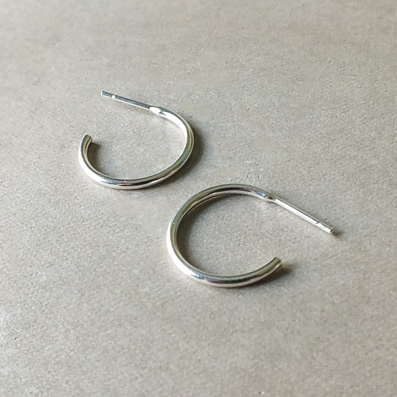 A pair of Open Hoop Earrings, medium from Becoming Jewelry, featuring a simple and classic silver design with open ends, are placed on a flat, light-colored surface. These dainty hoops are lightweight and perfect for everyday wear.