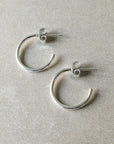 A pair of medium Open Hoop Earrings by Becoming Jewelry, lying on a textured beige surface, embodying lightweight classic elegance.