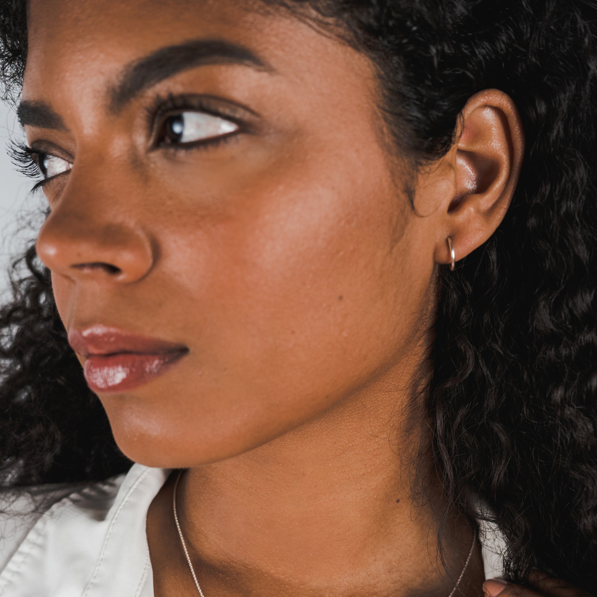 Side profile of a woman with curly hair, adorned with dainty Becoming Jewelry sterling silver open hoop earrings, small, looking away from the camera.