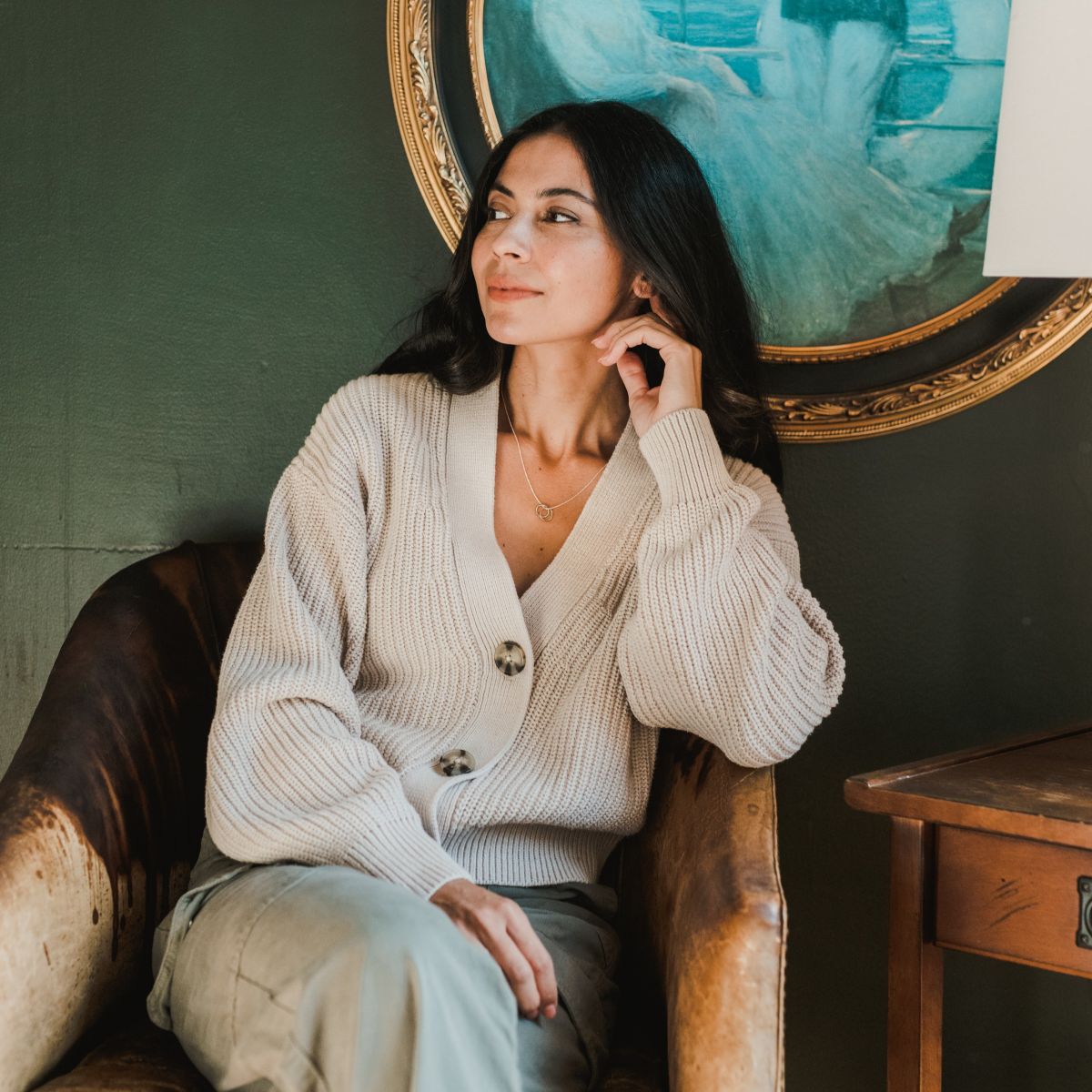 A woman wearing a beige cardigan is seated in a brown chair, gazing sideways. A captivating painting with textured blue circles adorns the green wall behind her, contrasting with her True Friends Necklace by Becoming Jewelry.